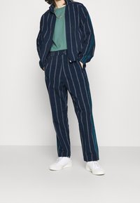 Navy track suit with vertical stripes in shades of blue and white. The jacket has a high collar and ribbed cuffs. Paired with a green top and white sneakers.