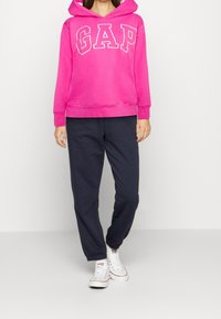 Sudadera de color rosa brillante con el logo "GAP" en blanco, combinada con pantalones de chándal azul marino oscuro. La sudadera tiene un bolsillo canguro y capucha con cordón ajustable.
