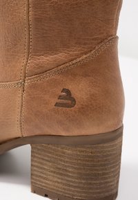 Hnědá kožená bota má texturovaný povrch, kontrastní prošívání a robustní dřevěný blokový podpatek, na kterém se nachází detail loga.