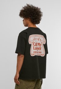 Sort bomulds t-shirt med korte ærmer, pryddet med et stort hvidt og orange grafisk design af en vintage telefon samt tekst på ryggen.