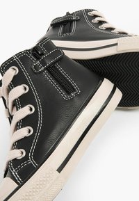 Sorte højhælede sneakers lavet af glat læder med kontrasterende hvid syning og snørebånd foran, med en lynlås på siden for nem påklædning.