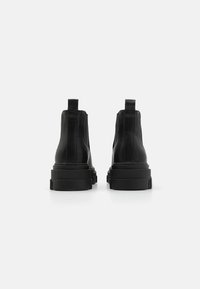 Botas de tobillo de cuero negro con suela gruesa, lengüetas en la parte trasera y detalles cosidos a lo largo de las costuras. Textura suave y diseño moderno.