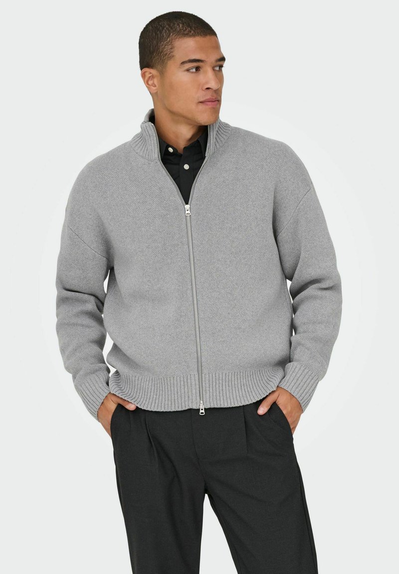 Pull gris en laine tricotée avec fermeture éclair, col haut, poignets et ourlet côtelés. Associé à un pantalon noir pour un look épuré et minimaliste.