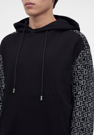 Jeune personne portant un sweat à capuche noir avec des manches à motifs comportant un texte blanc répétitif et des cordons avec des embouts argentés.