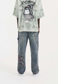 Graphic short-sleeve shirt in light green with a cartoon geisha design; loose blue denim pants with colorful embroidered accents on one leg.