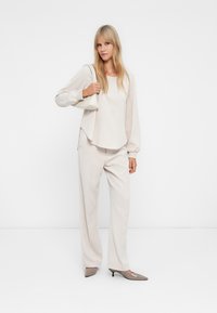 Γυναίκα που φοράει μπεζ φαρδιά μπλούζα και φαρδιές παν trousers, κρατώντας λευκή τσάντα ώμου, στέκεται μπροστά σε απλό λευκό φόντο.