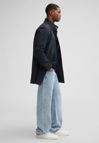 Men's black lightweight trench coat with a high collar, paired with loose light blue jeans and white sneakers. Simple design, no visible patterns.