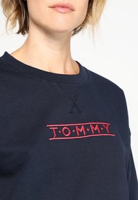 Felpa blu navy con scollo a girocollo e logo "TOMMY" ricamato in rosso sul petto. Tessuto morbido con dettagli di cucitura visibili.