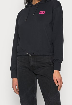 Person trägt einen schwarzen, kurzen Hoodie mit einem pinken FILA-Logo-Patch und schwarzen Jeans mit Kordelzug.