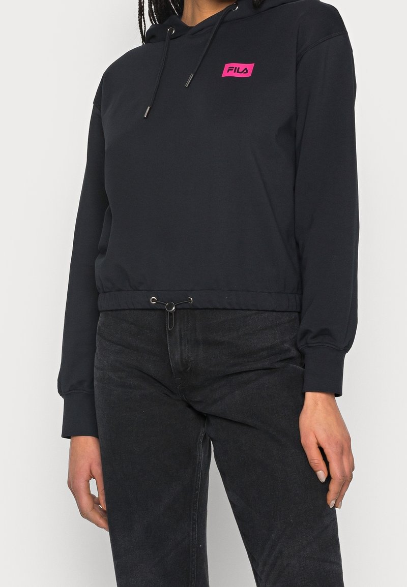 Personne portant un sweat à capuche court noir avec un patch logo FILA rose et un jean noir à cordon.