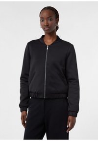 Frau mit geflochtenem Haar, die eine schwarze Kapuzenjacke und passende schwarze Hosen trägt, steht vor einem schlichten hellen Hintergrund.