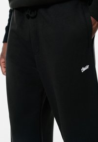 Schwarze Jogginghosen aus einem weichen Baumwoll-Mischgewebe, mit Seitentaschen und einem weißen Logo auf dem Oberschenkel. Elastischer Bund mit Kordelzug.