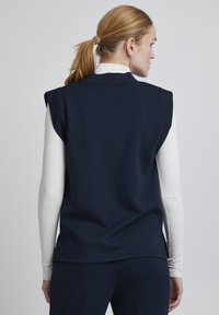 Top senza maniche blu navy con scollo rotondo e spacchi laterali, realizzato in tessuto morbido, indossato sopra una camicia a maniche lunghe bianca.