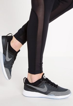 Jambes portant des leggings noirs avec panneaux en mesh et des chaussures de course Nike noires et blanches avec semelles blanches sur fond blanc.
