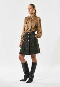 Blusa de satén bronce con detalle de lazo, falda a cuadros negra con acentos de botones dorados y botas de cuero negras hasta la rodilla. Entorno de estudio, fondo neutro.