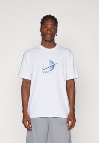 Λευκό βαμβακερό μπλουζάκι με κεντρικό μπλε γραφικό που περιέχει το λογότυπο της Nike και κυκλικά σχεδιαστικά στοιχεία. Έχει κοντά μανίκια και κωμική λαιμόκοψη.