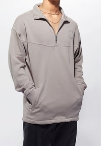 Světlounce šedý pulovr na čtvrtý zip z měkké látky, s vysokým límcem, žebrovanými manžetami a dvěma bočními kapsami. Jemný detail loga na hrudi.