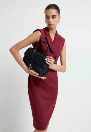 Mujer con un vestido sin mangas de color burdeos, sosteniendo un bolso de mimbre negro, llevando pendientes de aro dorados, de pie contra un fondo blanco liso.