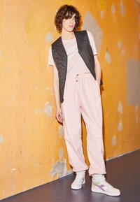 Ung person med lockigt hår iklädd en rosa Nike-träningsoverall och svart väst, stående mot en strukturmönstrad orange vägg och grått golv.