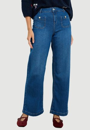 Frau trägt blaue Jeans mit weitem Bein, vorderen Klappentaschen und bordeauxfarbenen Mary Jane Schuhen, steht vor einfachem Hintergrund.
