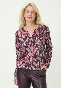 Bluse mit langen Ärmeln aus dunkelviolettem Stoff mit floralem Muster in Pink und Beige, V-Ausschnitt und weicher Textur. Kombiniert mit dunkler Lederhose.