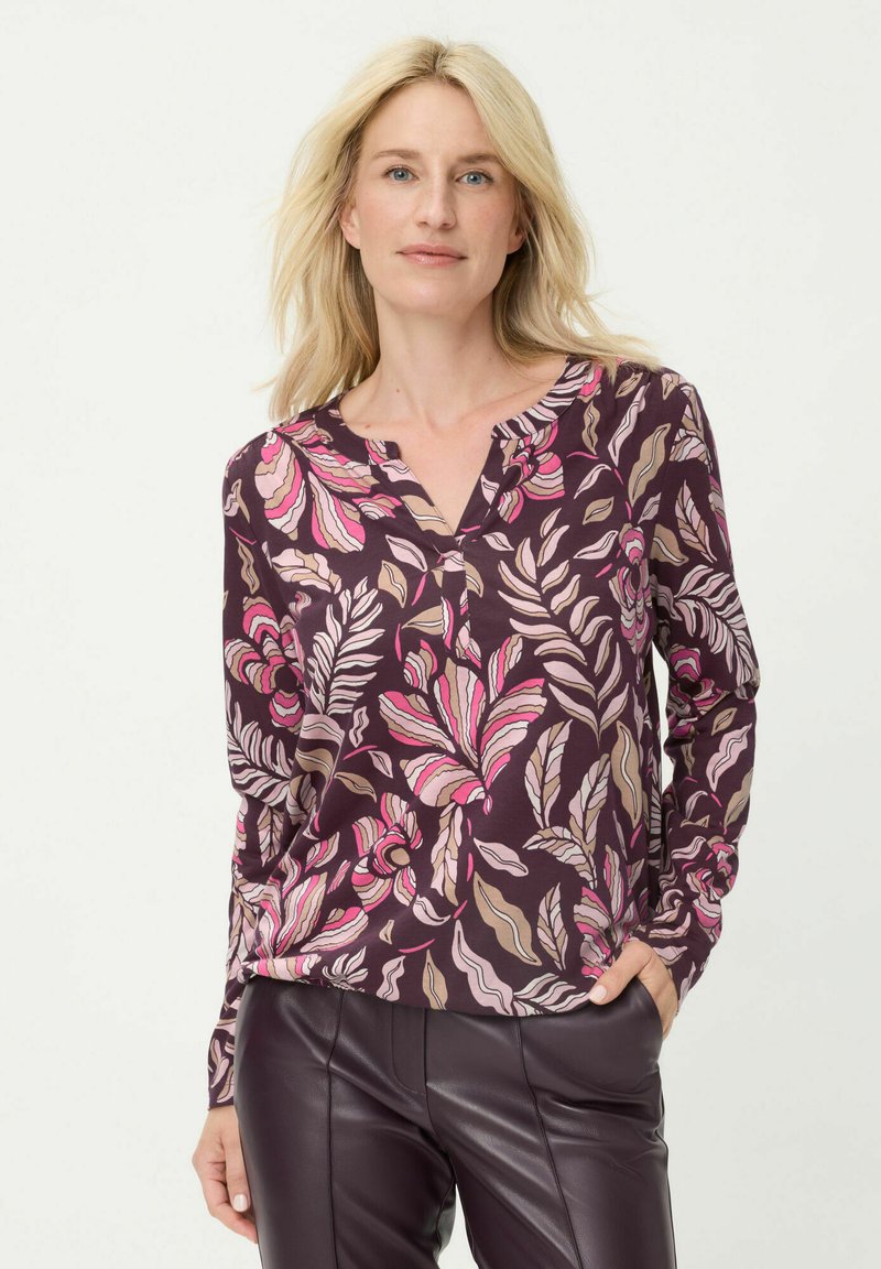 Bluse mit langen Ärmeln aus dunkelviolettem Stoff mit floralem Muster in Pink und Beige, V-Ausschnitt und weicher Textur. Kombiniert mit dunkler Lederhose.
