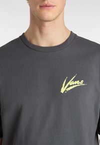 Camiseta de algodón gris con cuello redondo y mangas cortas, que presenta un logo de Vans en amarillo brillante impreso en el lado izquierdo del pecho.