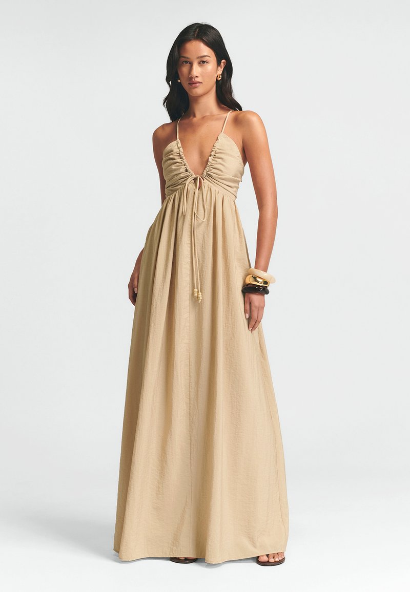 Maxi sukienka w beżowym kolorze, z głębokim dekoltem w kształcie litery V, marszczoną górą z troczkiem i zwiewnym materiałem. Gładka faktura i minimalne detale.