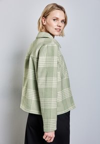 Zelená kostkovaná bunda s texturovaným materiálem, s límečkem, dlouhými rukávy a mírně zkráceným střihem. Jednoduché černé dno.