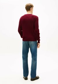 Pull côtelé bordeaux avec un décolleté en V, associé à un jean bleu délavé et des chaussures noires, vu de dos sur un fond blanc.