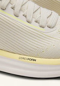 Közeli kép egy bézs színű kötött sportcipő felsőrészről, vastag, hozzáillő cipőfűzőkkel és világos sárga talppal, rajta "CORE x FOAM" párnázási technológia felirattal.