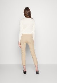 Krämfärgad kabelstickad tröja med lång ärm som kombineras med beige tajta byxor, sedd bakifrån. Svarta slip-on skor fulländar outfitten.