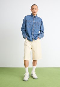Denim button-up shirt with two front pockets, paired with cream-colored knee-length shorts and white sneakers with gray accents.