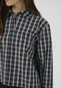 Sieviete, ģērbta melnbaltā pārbaudītā krekla ar garām piedurknēm un apkakli, redzama no kakla līdz viduklim uz neizteiksmīga fona.