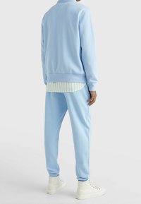 Ljusblå sweatshirt och byxor av mjukt tyg med avslappnad passform. Under den en grön- och vitrandig skjorta. Vita sneakers.