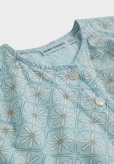 Chemise bébé bleu clair avec motifs de marguerites blanches en carrés, étiquetée "BOBO CHOSES", comportant deux boutons blancs près du col.
