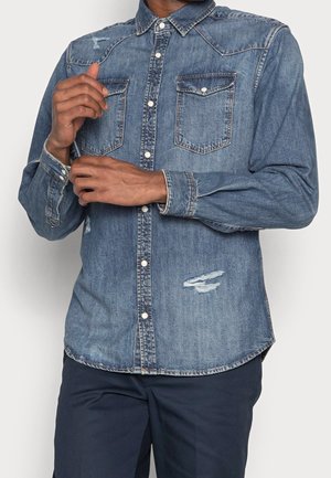 Homme portant une chemise en denim bleu délavé avec des poches poitrine à boutons et des détails usés, ajustant le poignet de sa chemise.