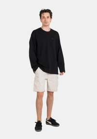 Camisa negra de manga larga con un logo en el frente, combinada con shorts cargo beige. El atuendo se complementa con zapatillas negras.