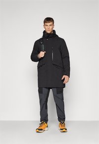 Palton negru izolat, cu buzunare cu fermoar, guler înalt și glugă, asociat cu pantaloni cargo gri și pantofi de drumeție portocalii. Textură netedă, design practic.