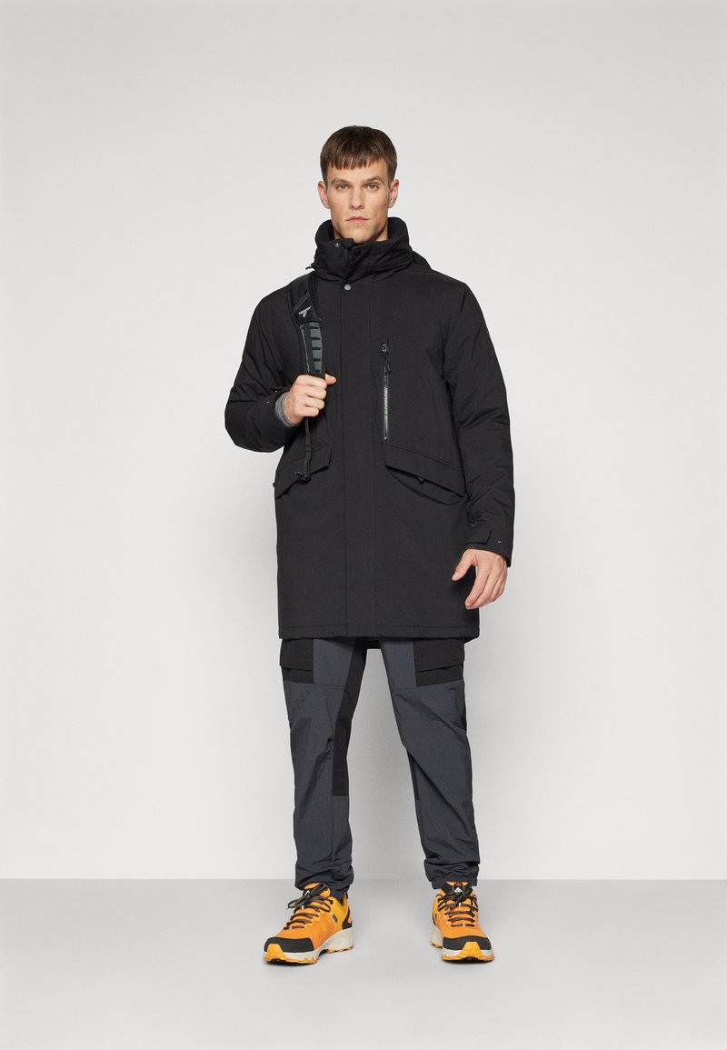 Czarna ocieplana kurtka z zamkami błyskawicznymi, wysoki kołnierz i kaptur, w połączeniu z szarymi spodniami cargo i pomarańczowymi butami turystycznymi. Gładka faktura, praktyczny design.