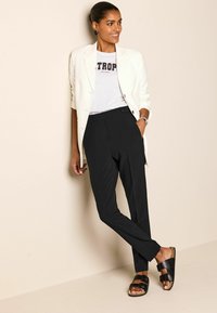 Weißer Blazer über einem grauen T-Shirt mit schwarzer Schrift, schwarze schmal geschnittene Hose und schwarze Sandalen mit Schnallen. Glatter Stoff und lässige Passform.