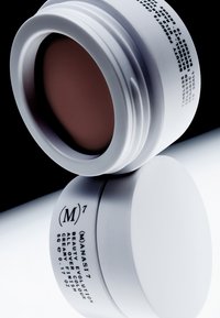 Kremowy, jasnobrązowy kosmetyk w białym, okrągłym opakowaniu z zakręcanym wieczkiem; na etykiecie znajduje się minimalistyczny czarny tekst.