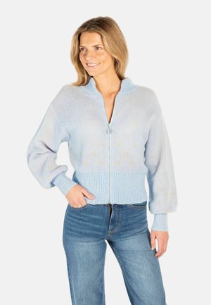 Smilende kvinne iført lys blå genser med glidelås, ribbestrikkede mansjetter og høy krage, kombinert med høytlivs blå jeans, stående med hånden i lommen.