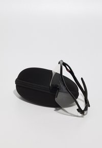 Schwarze Sportsonnenbrille mit transparenten Gläsern, die auf einem schwarzen ovalen Hartschalenetui mit Reißverschluss und matter Oberfläche liegt.
