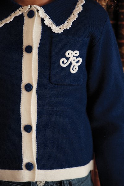 Cardigan azul-marinho com botões e fechos brancos, apresentando um decote com colarinho de rendas e um bolso frontal com um logótipo bordado em branco.