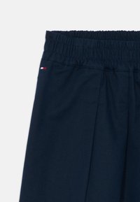 Marineblauwe broek met een elastische tailleband, voorzien van plooien en een klein logo etiket in rood, wit en blauw aan de zijkant. Gladde katoen stof.