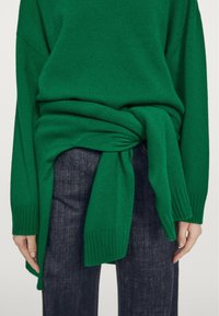 Grüner Strickpullover, in der Taille gebunden, mit gerippten Bündchen und lässiger Passform, kombiniert mit dunklen Denim-Jeans.