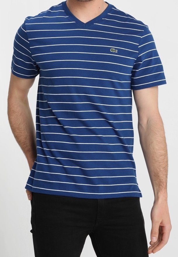 Man in een blauwe V-hals T-shirt met dunne witte horizontale strepen en zwarte broek tegen een effen achtergrond.