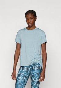 Lys blå kortærmet T-shirt med rund halsudskæring og rynkede detaljer på den ene side. Sammen med mønstrede blå og brune leggings.