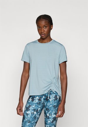 T-shirt azul claro de manga curta com decote redondo e detalhes franzidos de um lado. Combinado com leggings estampadas em azul e castanho.